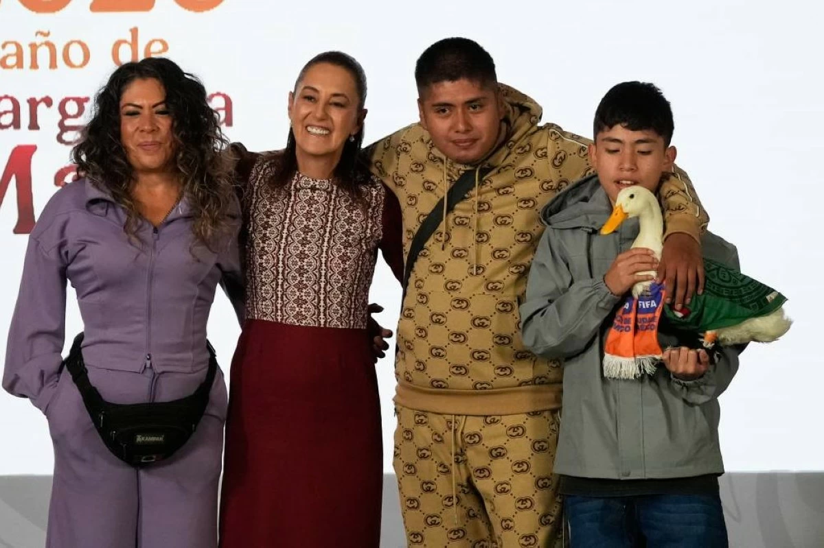 Merlin the duck, dressed in a Mexican national soccer team jersey, attends the daily press conference by Mexican President Claudia Sheinbaum, second from left, along with his caretakers Carla Gomez and her sons Carlos, and Christian who holds Merlin, at the National Palace in Mexico City, Monday, June 22, 2026. (AP Photo/Marco Ugarte)